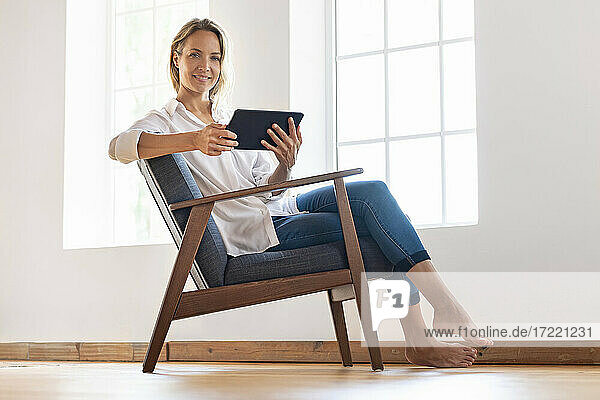 Lächelnde Frau mit digitalem Tablet auf einem Stuhl zu Hause