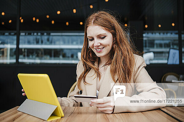 Lächelnde Frau schaut auf ihre Kreditkarte  während sie in einem Café vor einem digitalen Tablet sitzt