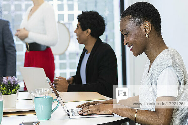 Smiling businesswoman working on laptop with colleagues in background at office