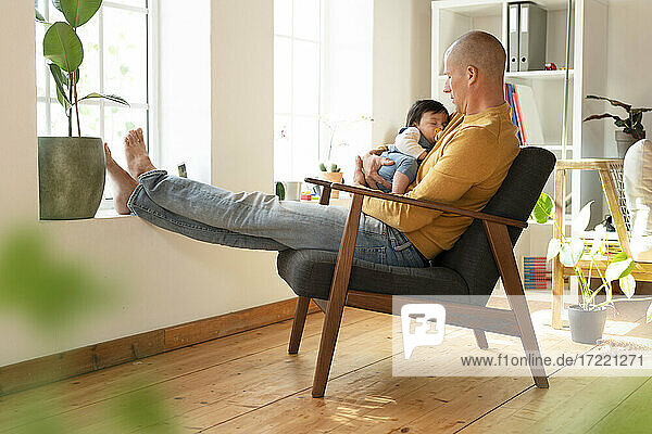 Vater sieht sein schlafendes Baby an  während er zu Hause auf einem Sessel sitzt