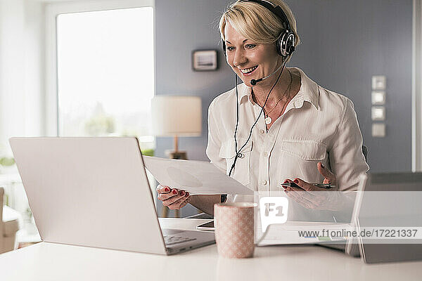 Female professional with paper talking on video call through laptop at home office