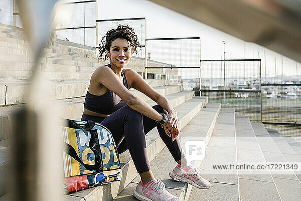 Lächelnde Sportlerin  die mit einer Tasche auf einer Treppe sitzt und träumt