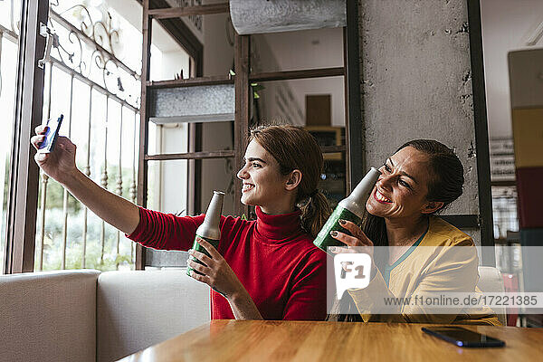 Cheerful female friends with beer bottles taking selfie through mobile phone in bar