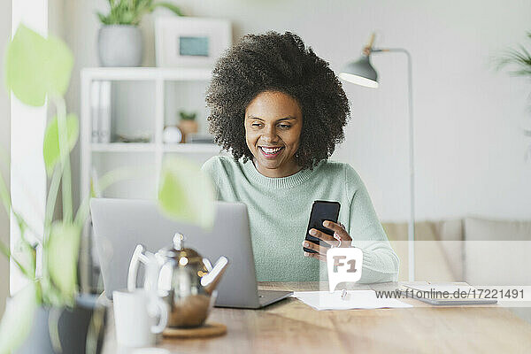 Smiling woman with mobile phone using laptop at home