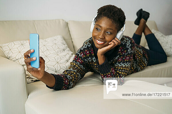 Smiling woman talking selfie through smart phone while listening music on sofa at home