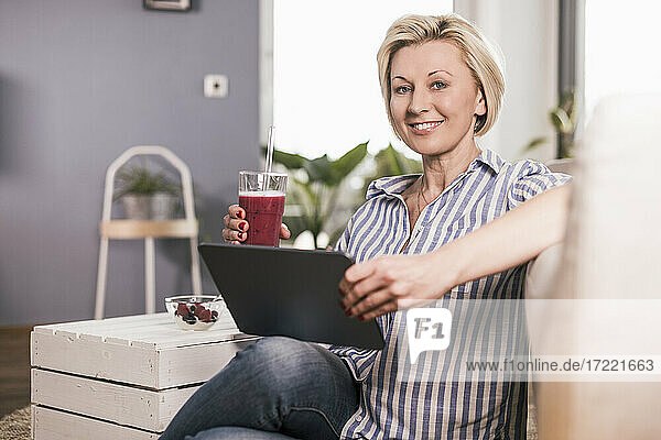 Lächelnde Frau  die ein digitales Tablet und ein Saftglas hält  während sie zu Hause sitzt
