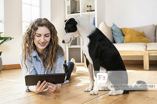 Smiling woman looking at digital tablet while lying on hardwood floor by Jack Russell Terrier in living room