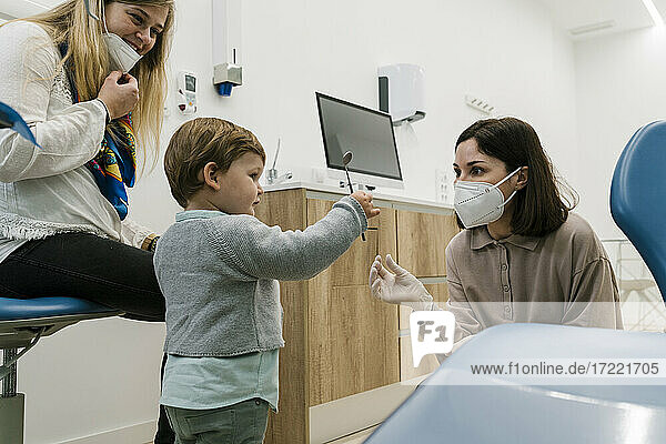 Junge spielt mit zahnmedizinischen Geräten  während er neben dem Zahnarzt und der Mutter steht  die in der Klinik eine Gesichtsmaske trägt