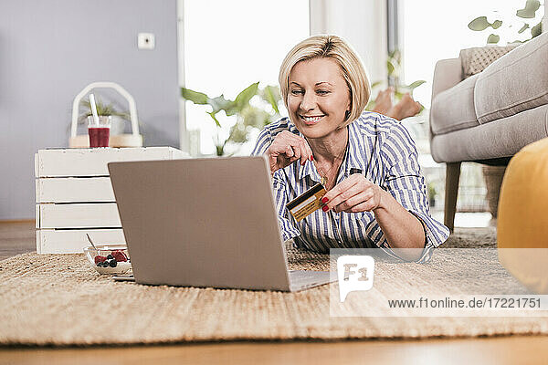Blonde reife Frau mit Kreditkarte beim Online-Shopping mit Laptop zu Hause