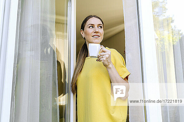Lächelnde Frau schaut weg und hält eine Kaffeetasse am Fenster