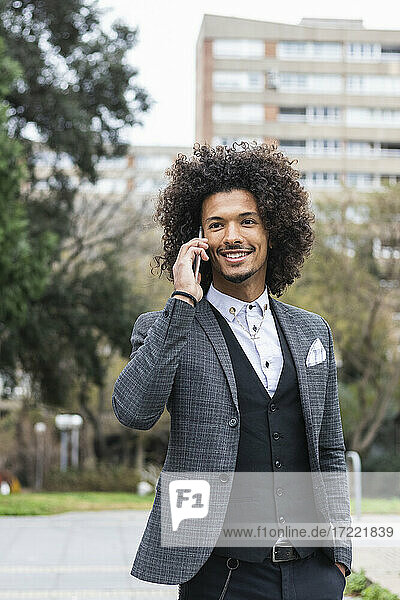 Lächelnder Geschäftsmann mit Hand in der Tasche  der mit einem Smartphone in der Stadt spricht
