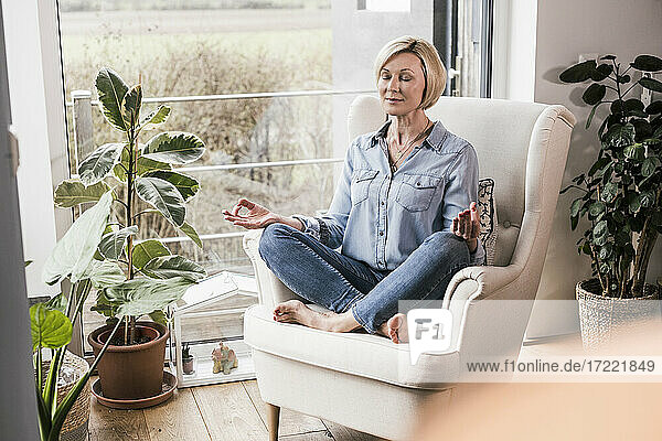 Reife Frau meditiert auf einem Sessel im Wohnzimmer sitzend
