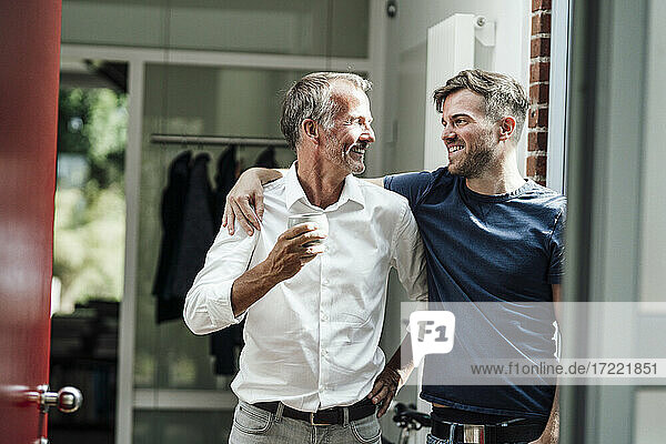 Happy son with arm around father holding coffee cup at home
