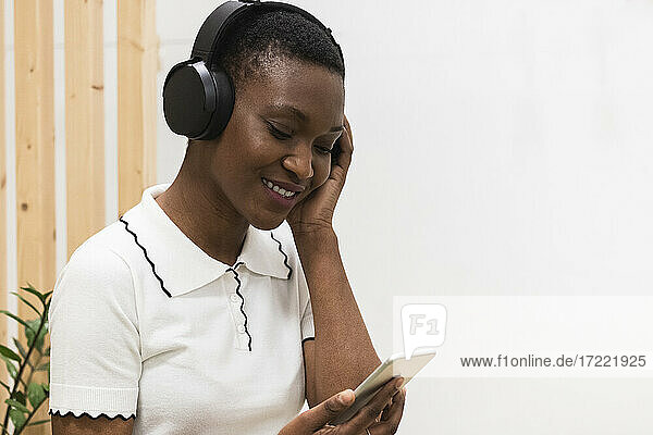 Lächelnde Geschäftsfrau hört Musik über Kopfhörer  während sie ein Mobiltelefon benutzt