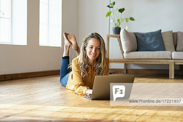 Lächelnde Frau  die einen Laptop benutzt  während sie auf dem Hartholzboden im Wohnzimmer liegt
