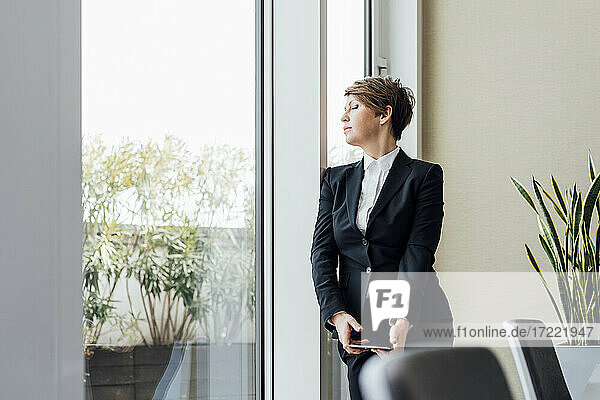 Female business professional with eyes closed holding digital tablet while standing by window in office