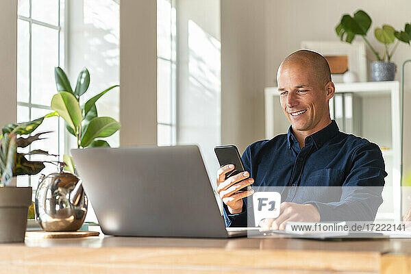 Smiling businessman with laptop using mobile phone while working at home