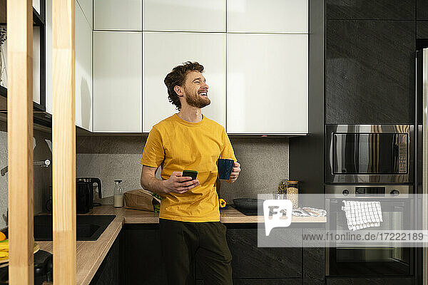 Glücklicher Mann mit Kaffeetasse und Smartphone in der heimischen Küche