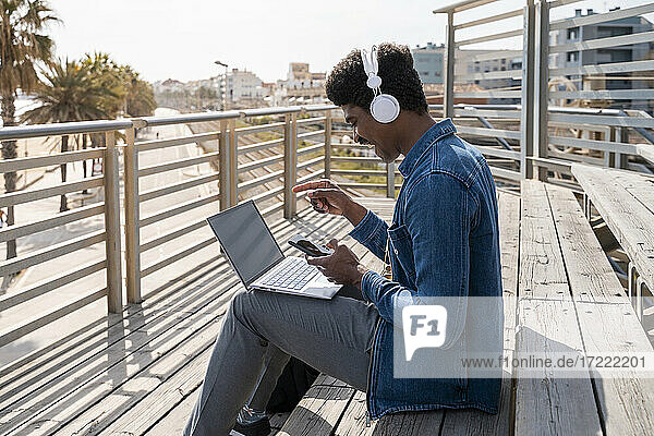 Fröhlicher Mann  der mit seinem Smartphone auf einen Laptop zeigt  während er auf einer Holztreppe an der Promenade sitzt
