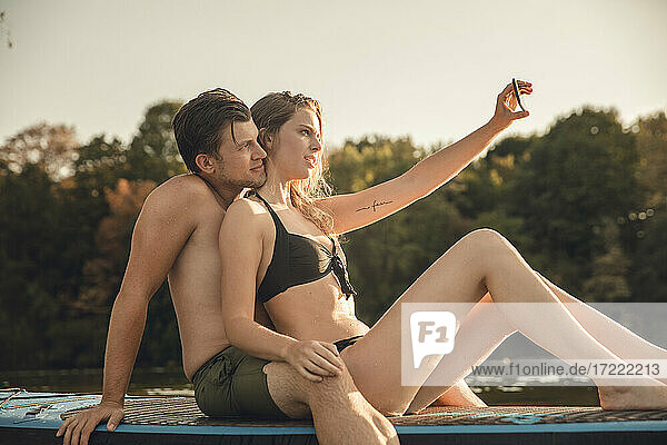 Glückliches Paar sitzt auf einem Paddelbrett auf einem See und macht Smartphone-Selfies