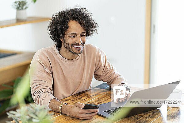 Lächelnder Geschäftsmann  der sein Smartphone hält  während er zu Hause am Laptop arbeitet