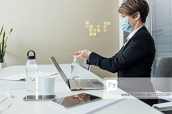 Weibliche Unternehmerin mit Gesichtsschutzmaske  die am Schreibtisch sitzend Händedesinfektionsmittel benutzt