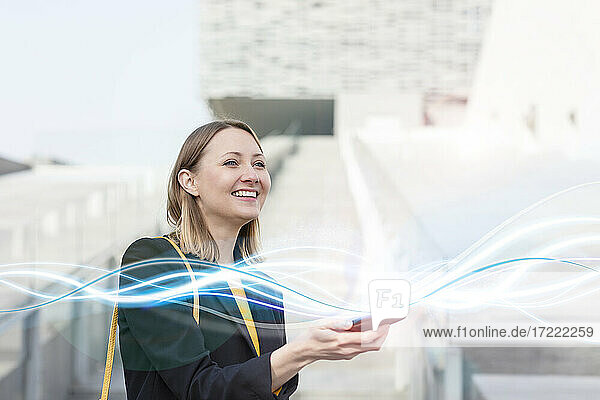 Lächelnde Unternehmerin  die ein leuchtendes Wellenmuster mit einem Mobiltelefon im Freien betrachtet