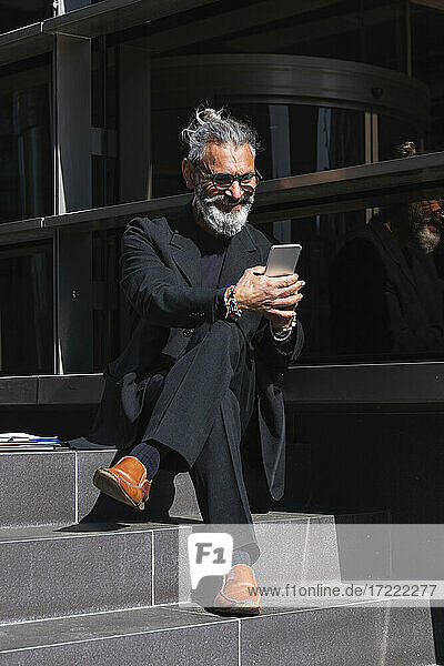 Lächelnder männlicher Berufstätiger  der ein Mobiltelefon benutzt  während er auf den Stufen eines Gebäudes sitzt