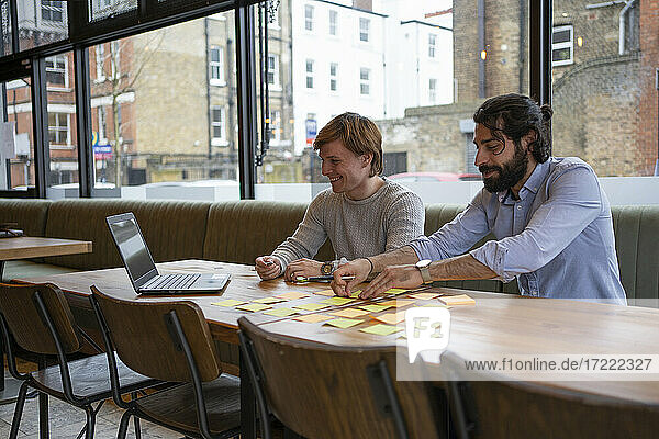 Smiling male entrepreneurs sticking adhesive note on desk while working over laptop in office