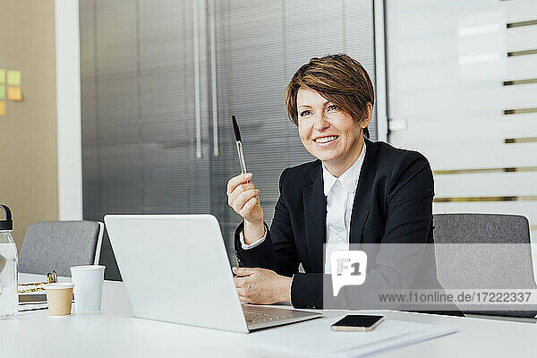 Lächelnde Unternehmerin hält einen Stift vor einem Laptop am Schreibtisch im Büro