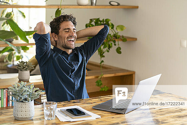 Glücklicher männlicher Freiberufler mit Händen hinter dem Kopf lachend  während er zu Hause vor seinem Laptop sitzt