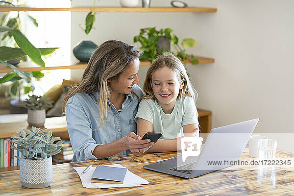 Businesswoman and daughter sitting with laptop and smart phone at table