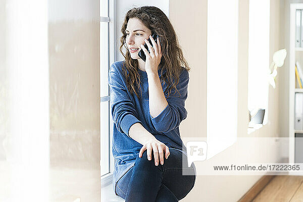 Junge Frau schaut durch das Fenster  während sie zu Hause mit dem Handy telefoniert