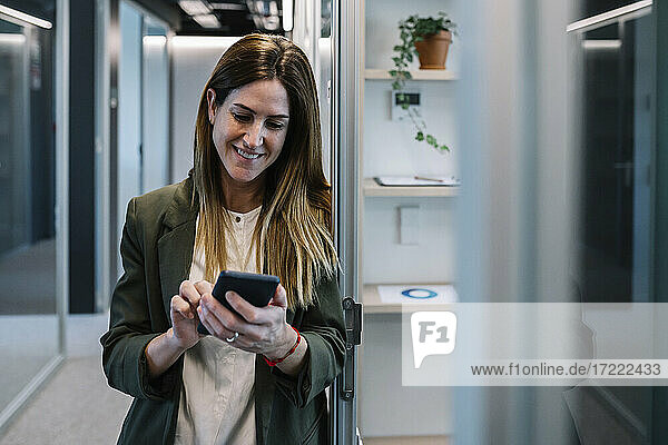 Smiling woman using smart phone while leaning on wall in corridor at office