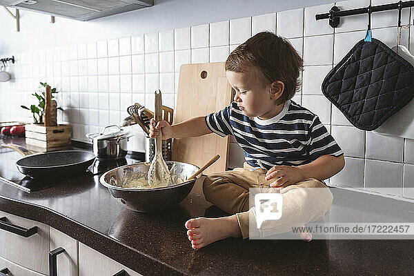 Cute boy mixing ingredients in bowl while sitting on kitchen counter at home