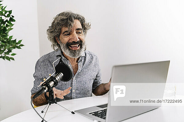 Smiling male podcaster sitting in front of laptop on table at office