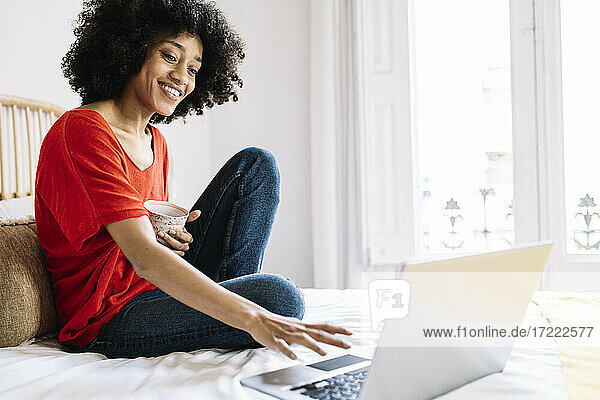 Junge Frau mit Tasse  die einen Laptop benutzt  während sie zu Hause auf dem Bett sitzt