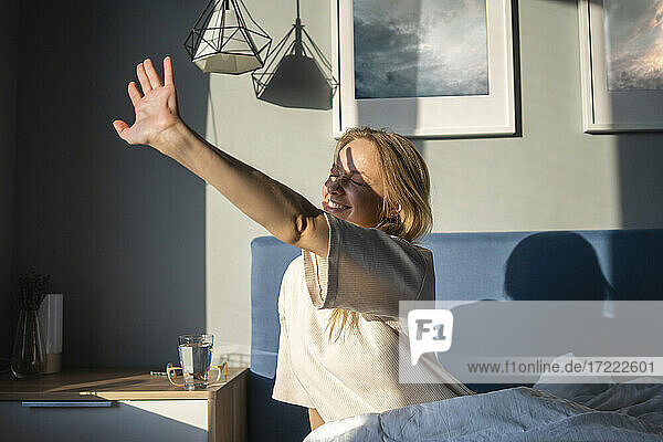 Lächelnde Frau streckt ihre Hand dem Sonnenlicht entgegen  während sie zu Hause auf dem Bett sitzt