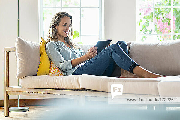 Smiling woman in casuals looking at digital tablet while sitting on couch