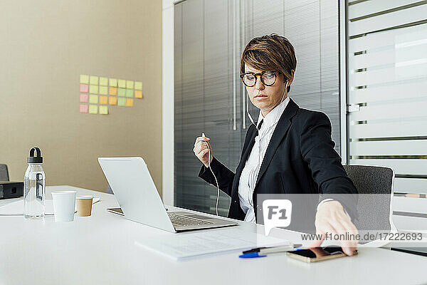 Geschäftsfrau mit In-Ear-Kopfhörern  die ein Mobiltelefon am Schreibtisch hält