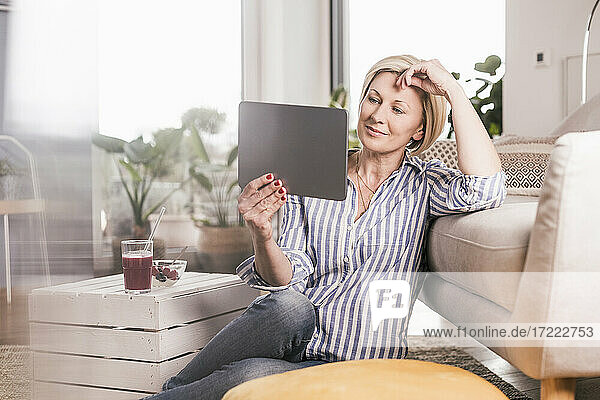 Ältere Frau schaut auf ein digitales Tablet  während sie sich zu Hause am Sofa anlehnt