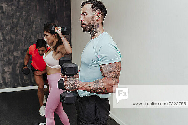 Sportler hält eine Hantel  während er mit Freunden im Hintergrund im Fitnessstudio trainiert