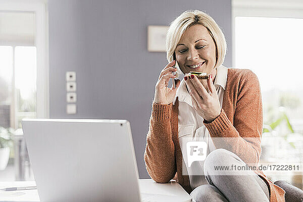 Lächelnde Geschäftsfrau mit geschlossenen Augen  die Brot isst  während sie mit dem Handy im Büro zu Hause telefoniert