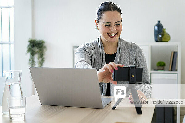 Glückliche Geschäftsfrau  die ihr Smartphone während eines Videogesprächs zu Hause auf einem Stativ einstellt