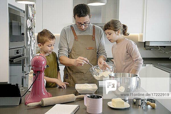Vater hält Schüssel mit Mehl und steht mit Kindern in der Küche