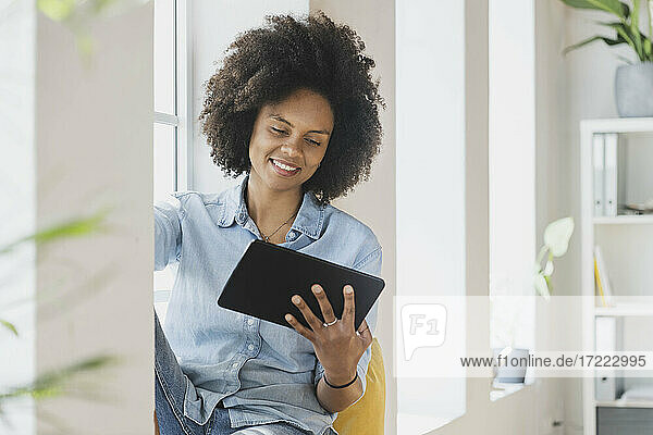 Lächelnde Frau  die ein digitales Tablet benutzt  während sie am Fenster sitzt