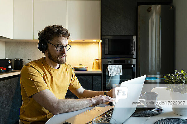 Male freelance worker listening through headphones while using laptop in kitchen