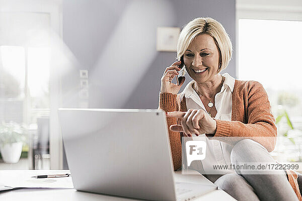 Lächelnde weibliche Fachkraft  die auf einen Laptop zeigt  während sie über ein Smartphone zu Hause kommuniziert