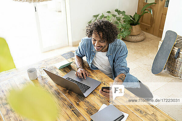 Geschäftsmann  der ein Smartphone hält  während er einen Laptop auf einem Tisch zu Hause benutzt