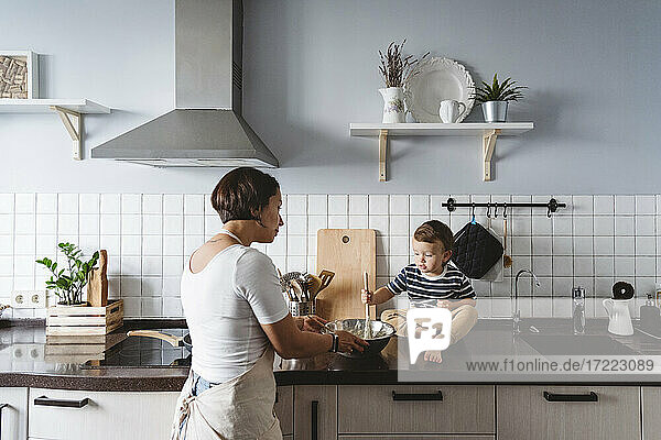Mother looking at cute child playing with bowl sitting on kitchen counter at home